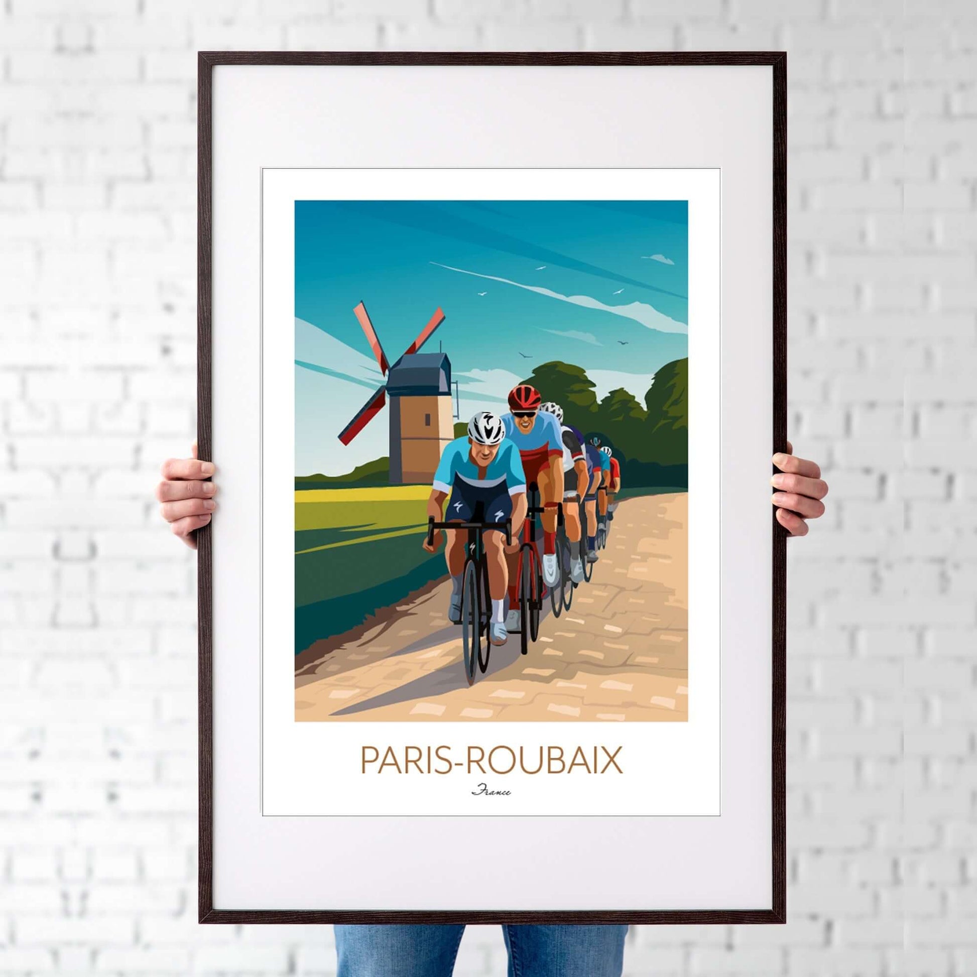 Man holding an A1 framed art print of the Paris Roubaix cyclists racing across cobble stone lane with an old traditional windmill in the background..