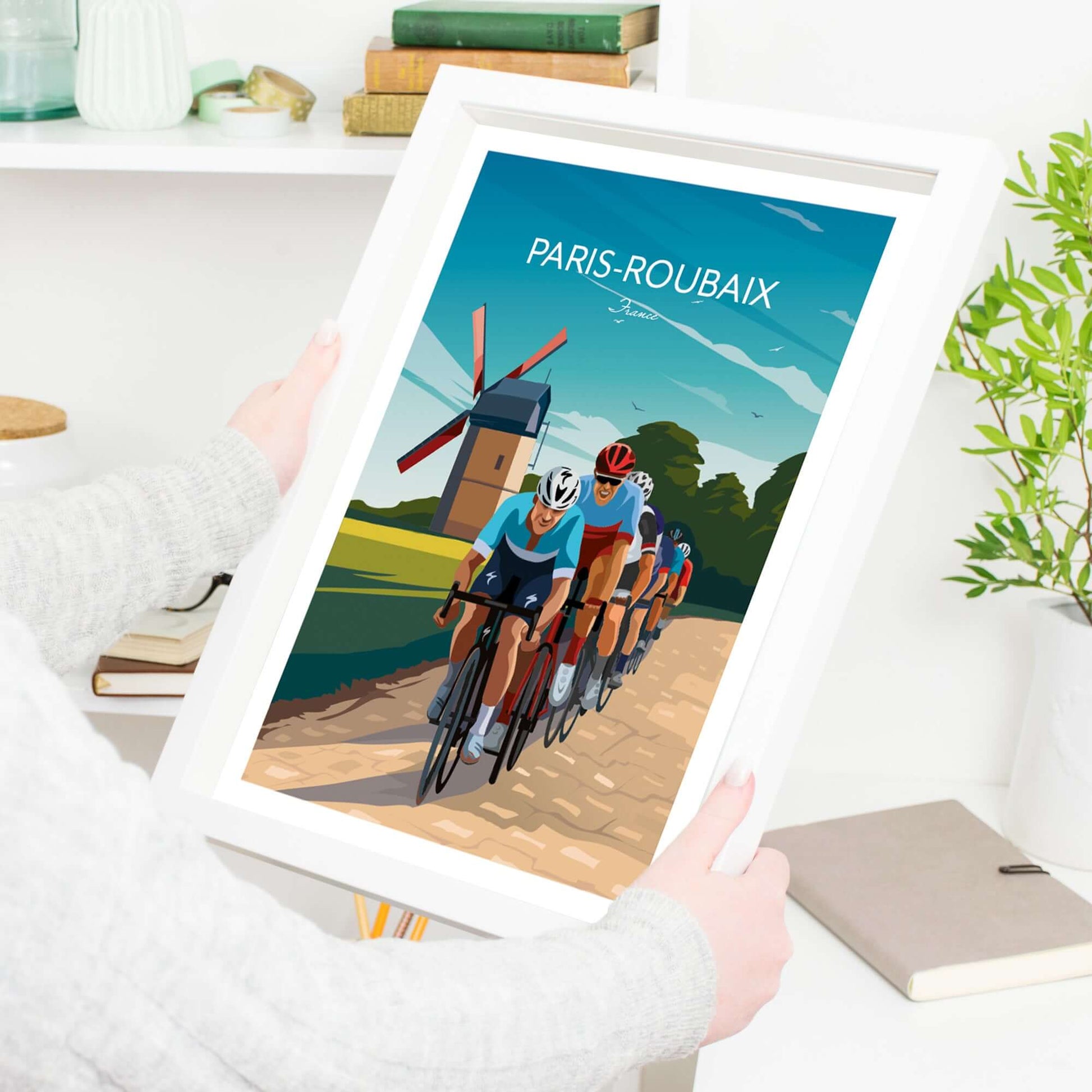 Vibrant cycling print of the Paris Roubaix in a white frame being held by a lady, ready to be put on display in a home interior.