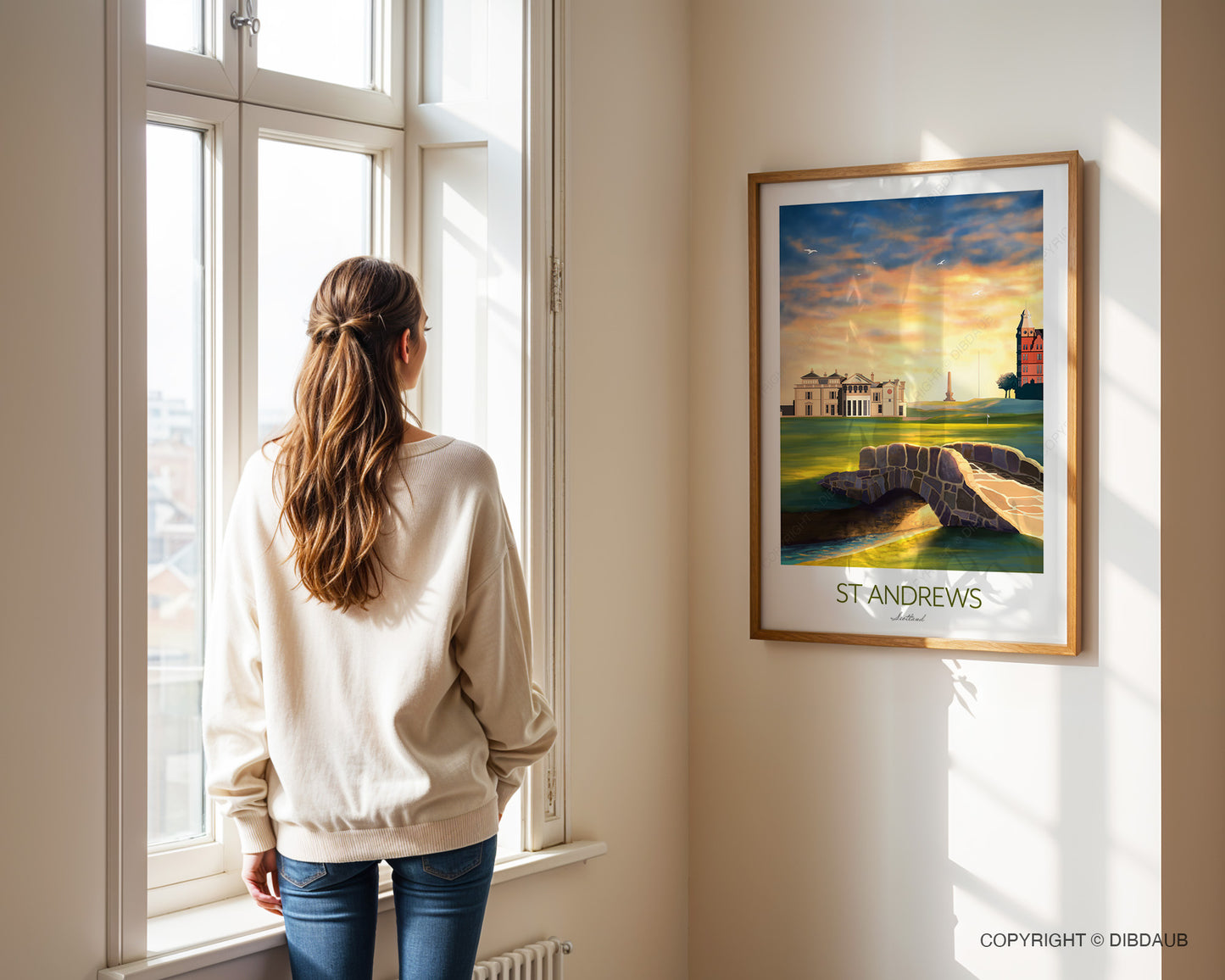 Woman standing by a window looking at a framed picture of St. Andrews on the wall.