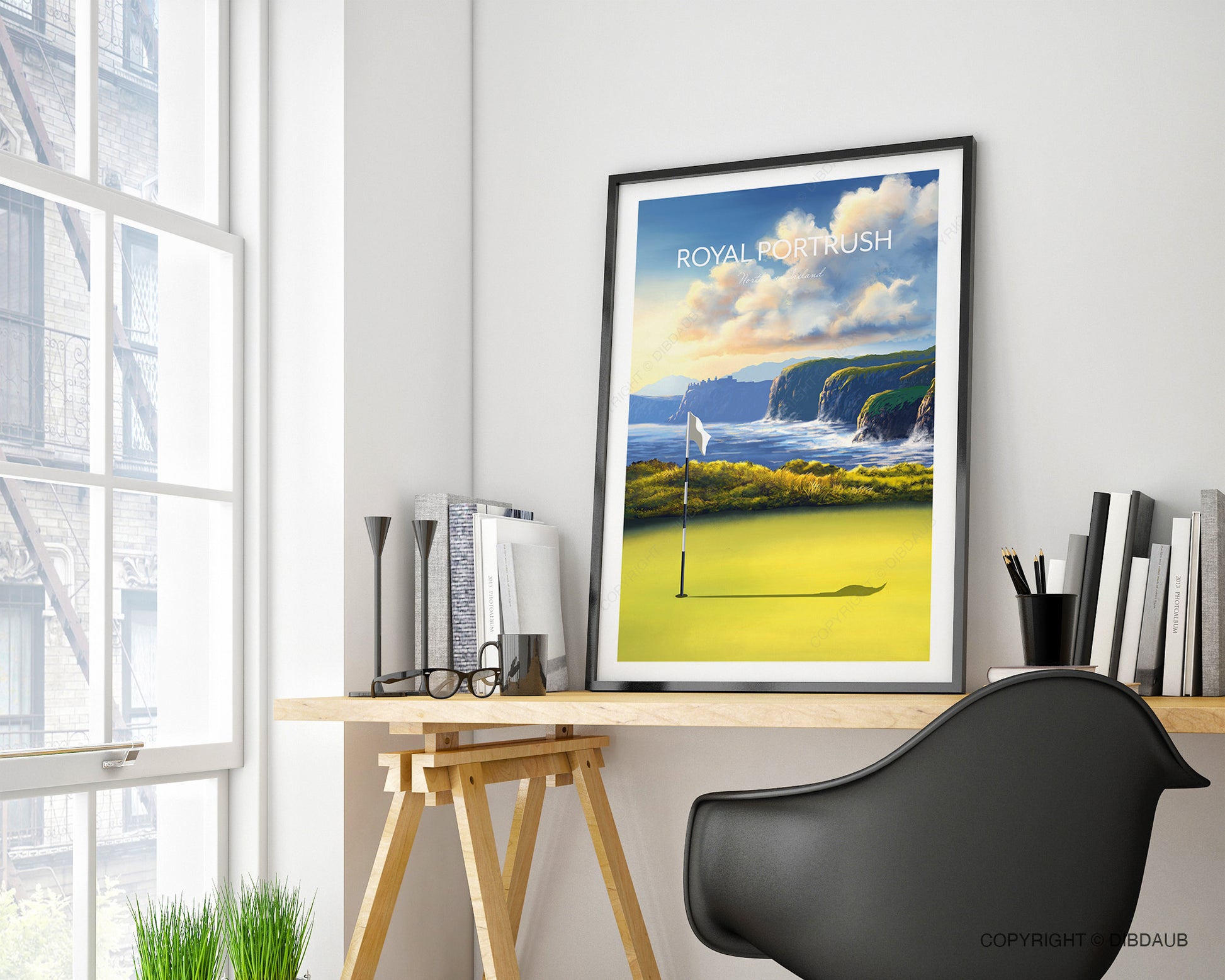 Framed artwork of a golf course with cliffs on a home office desk near a window.