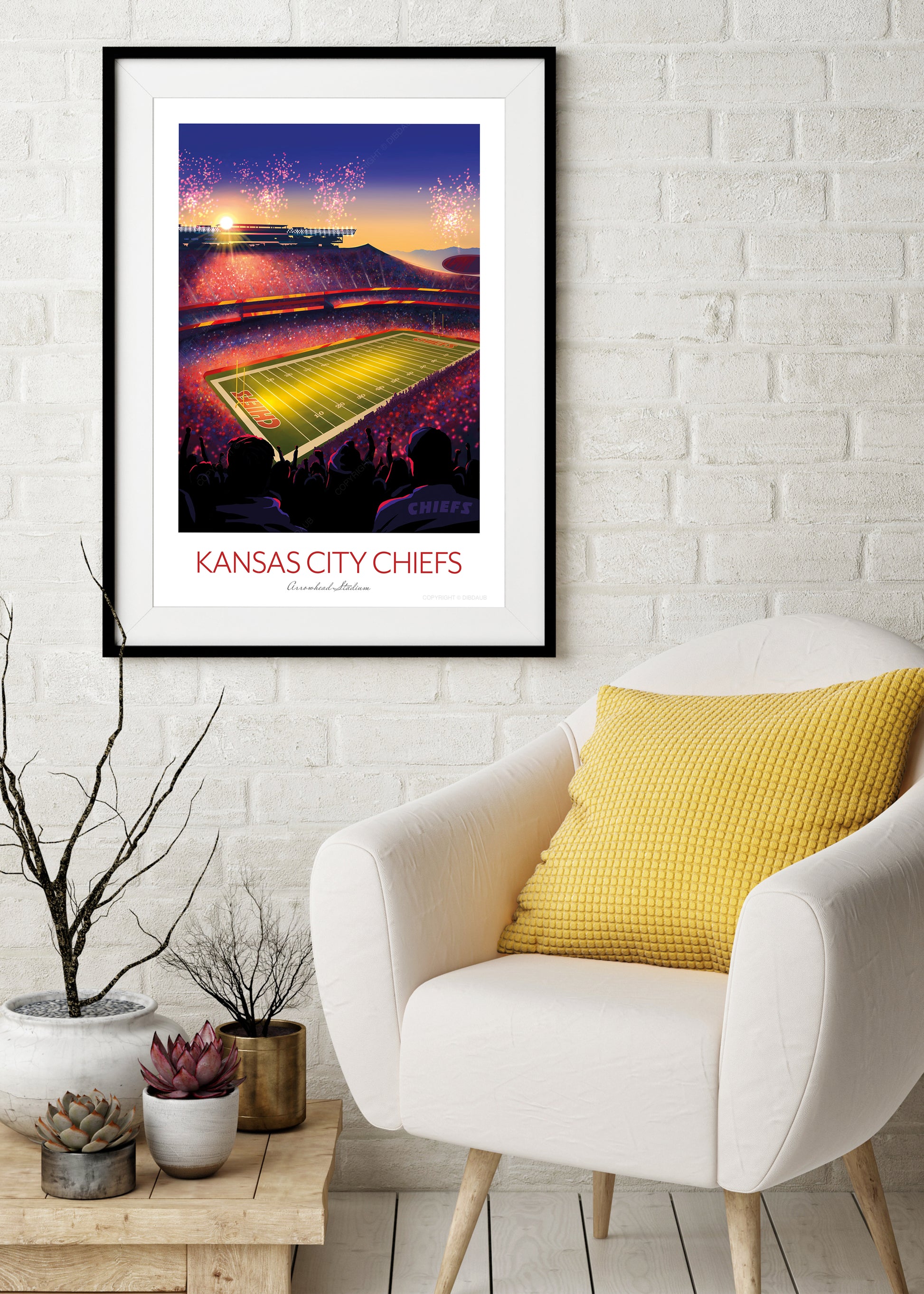Framed artwork of a football stadium with 'Kansas City Chiefs' text on a white brick wall above a white armchair with a yellow pillow.