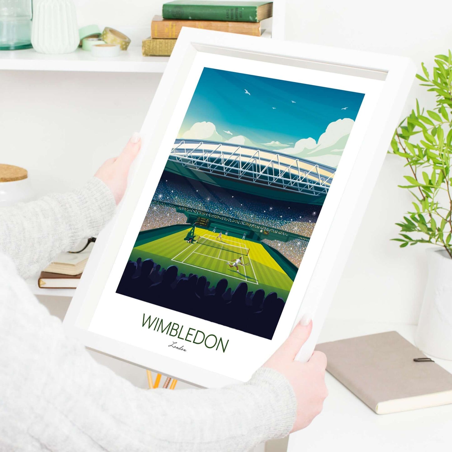 Tennis art print of Wimbledon in a white frame, being held by a female in a study with books and a plant in the background.