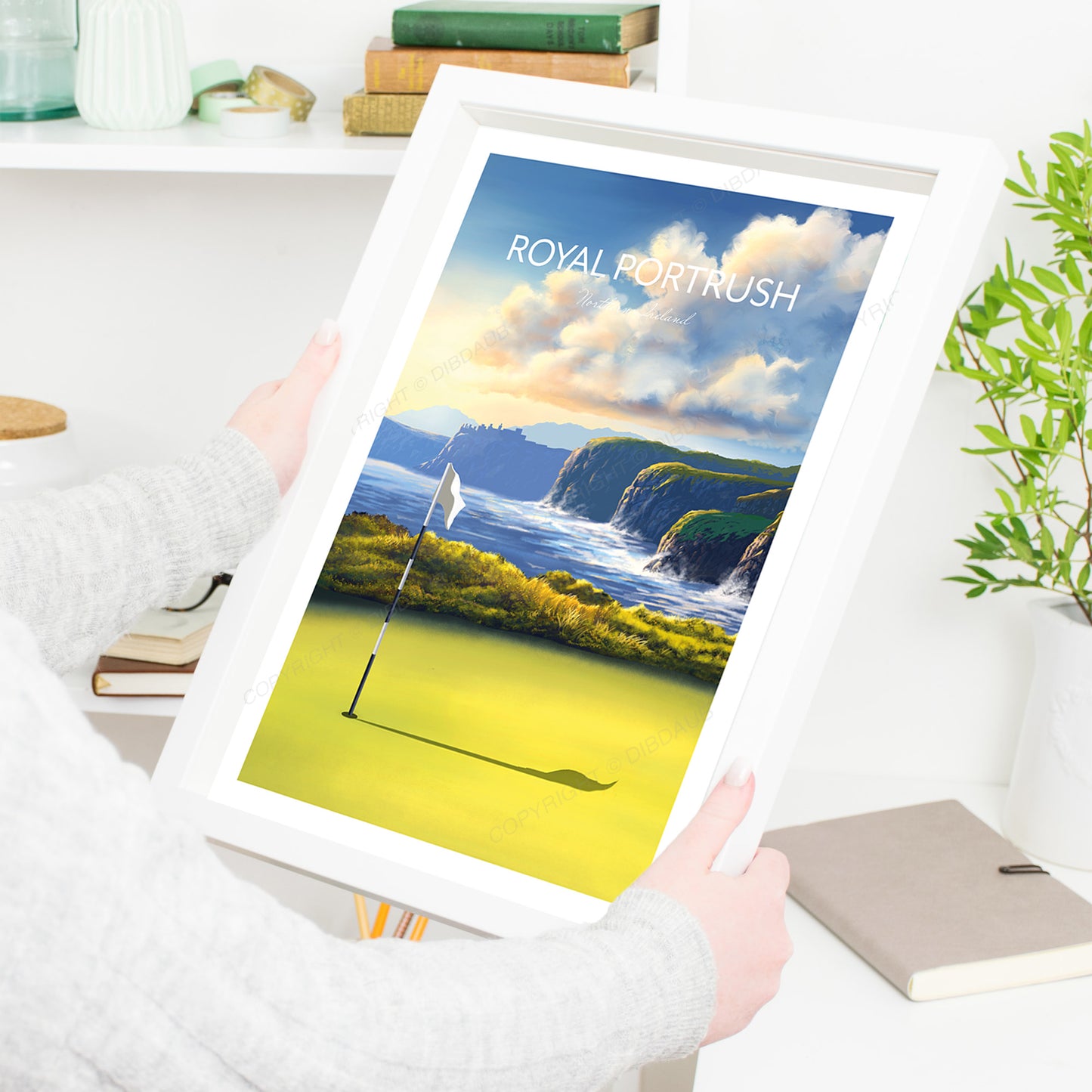 Framed artwork of Royal Portrush golf course in a white frame, being held by a person in a living room.