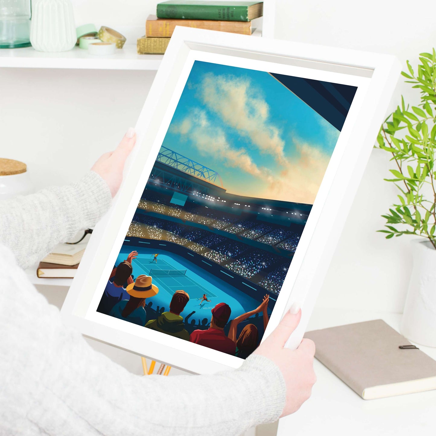 A lady holding a white framed tennis print of the Australian Open, with tennis players playing on centre court and fans watching.