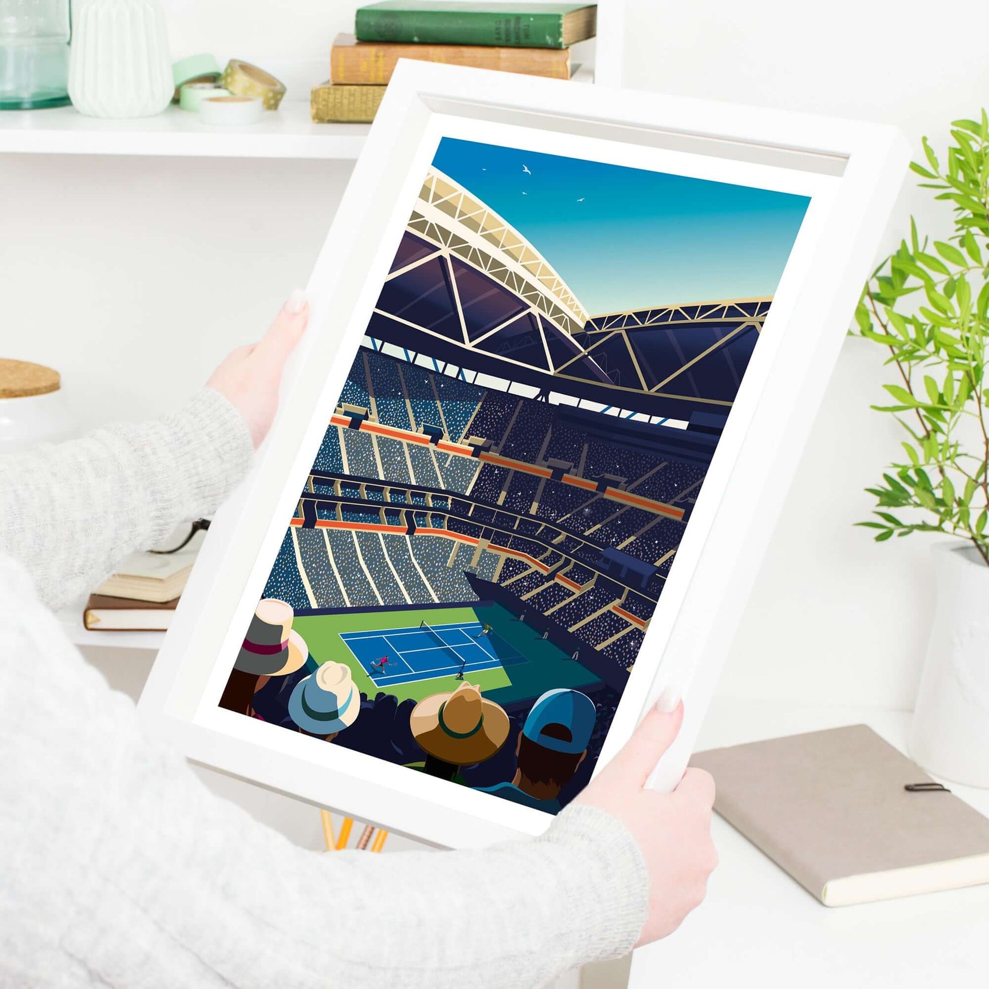 Lady holding a framed Tennis art print of Arthur Ashe Stadium, with tennis players in action in the US Open and fans viewing from a distance..