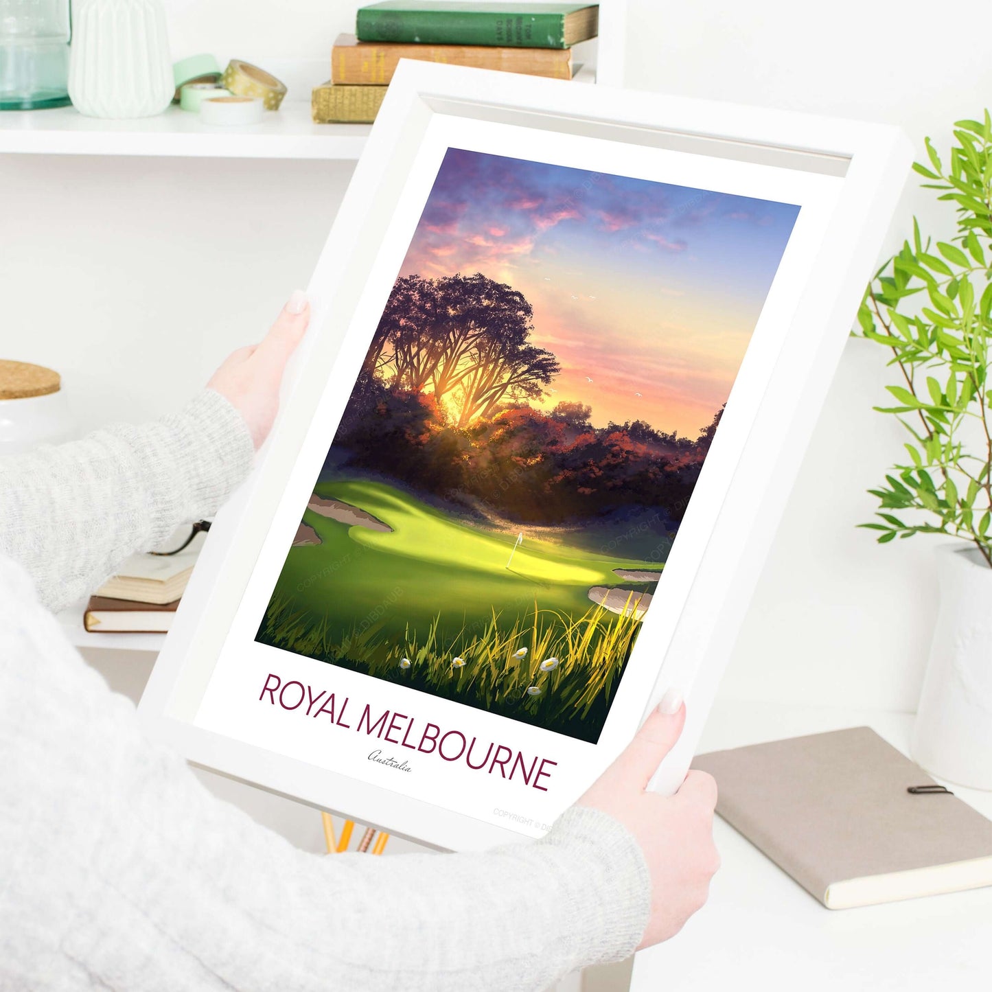 A lady holding a framed golf print of Royal Melbourne in a living room.